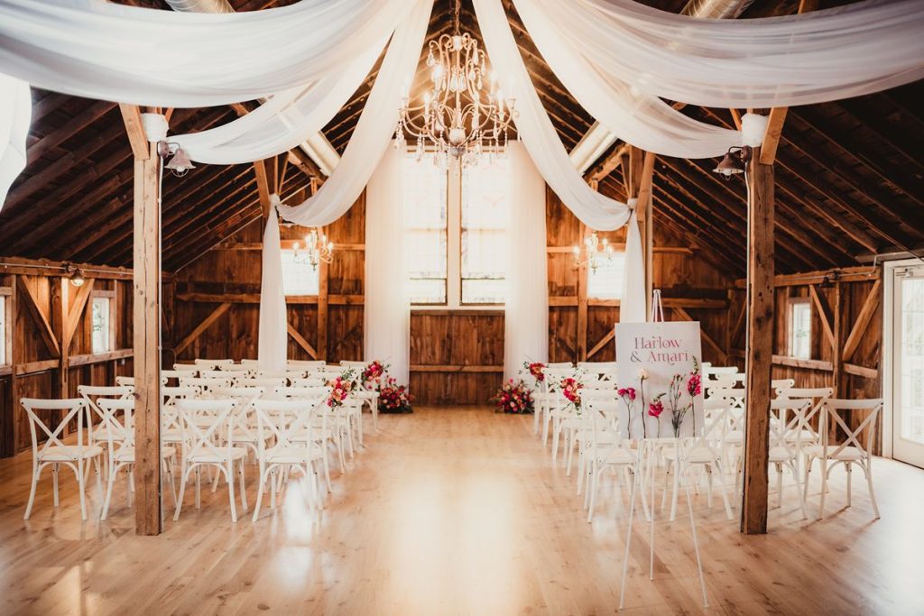 Indoor barn ceremony space with white chairs, draping fabric and pink flowers.